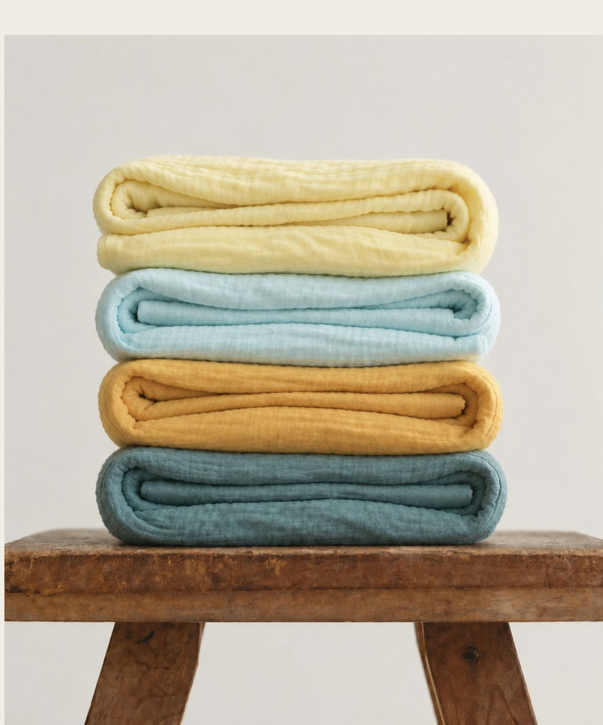 Stack of folded White Flowerr muslin towels in yellow, sky blue, mustard and teal on a wooden stool