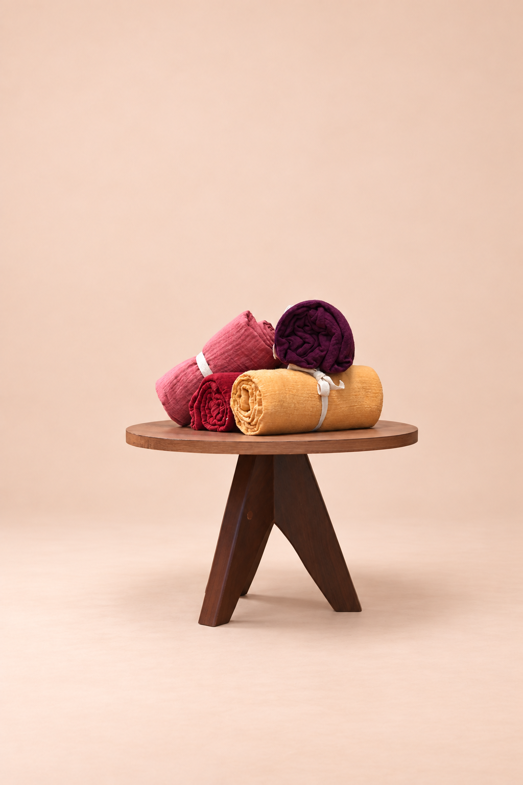 Rolled cotton muslin towels in pink, burgundy, purple and yellow on a wooden stool against a beige studio background
