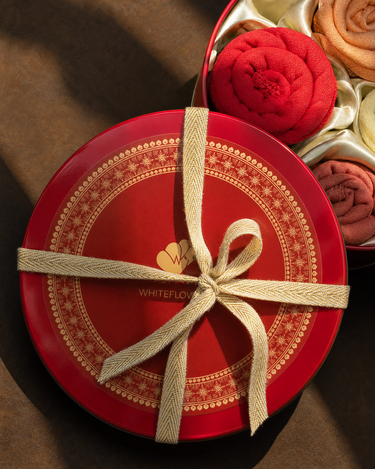 A round red gift box with gold decorative patterns and a cream ribbon tied in a bow, shown in a styled product shot. Part of the box contains white flower-shaped confectionery items wrapped in cream satin, including a prominent red flower-shaped piece.